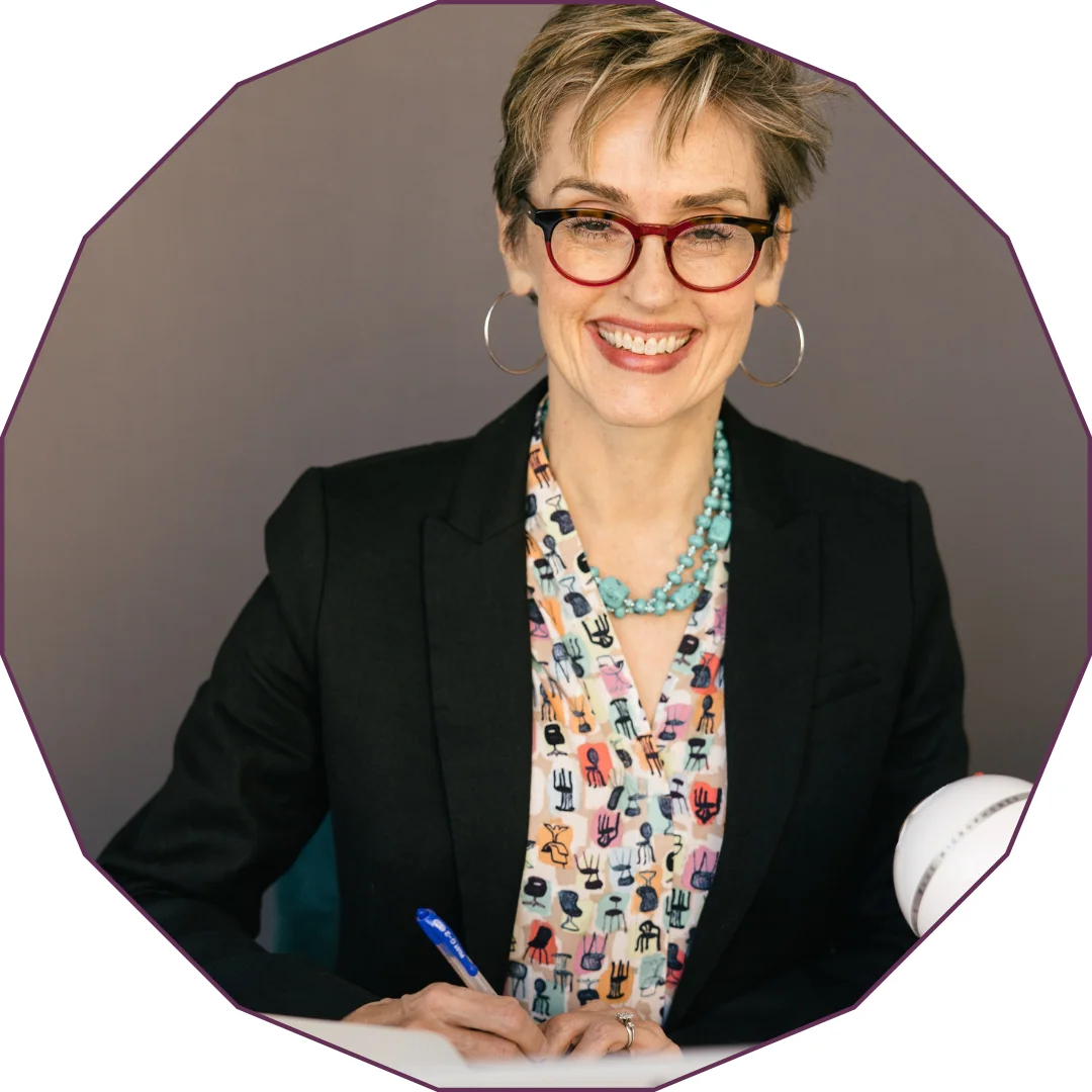 Smiling woman with short blonde hair wearing red-framed glasses, black blazer, turquoise beaded necklace, and patterned blouse, holding a pen