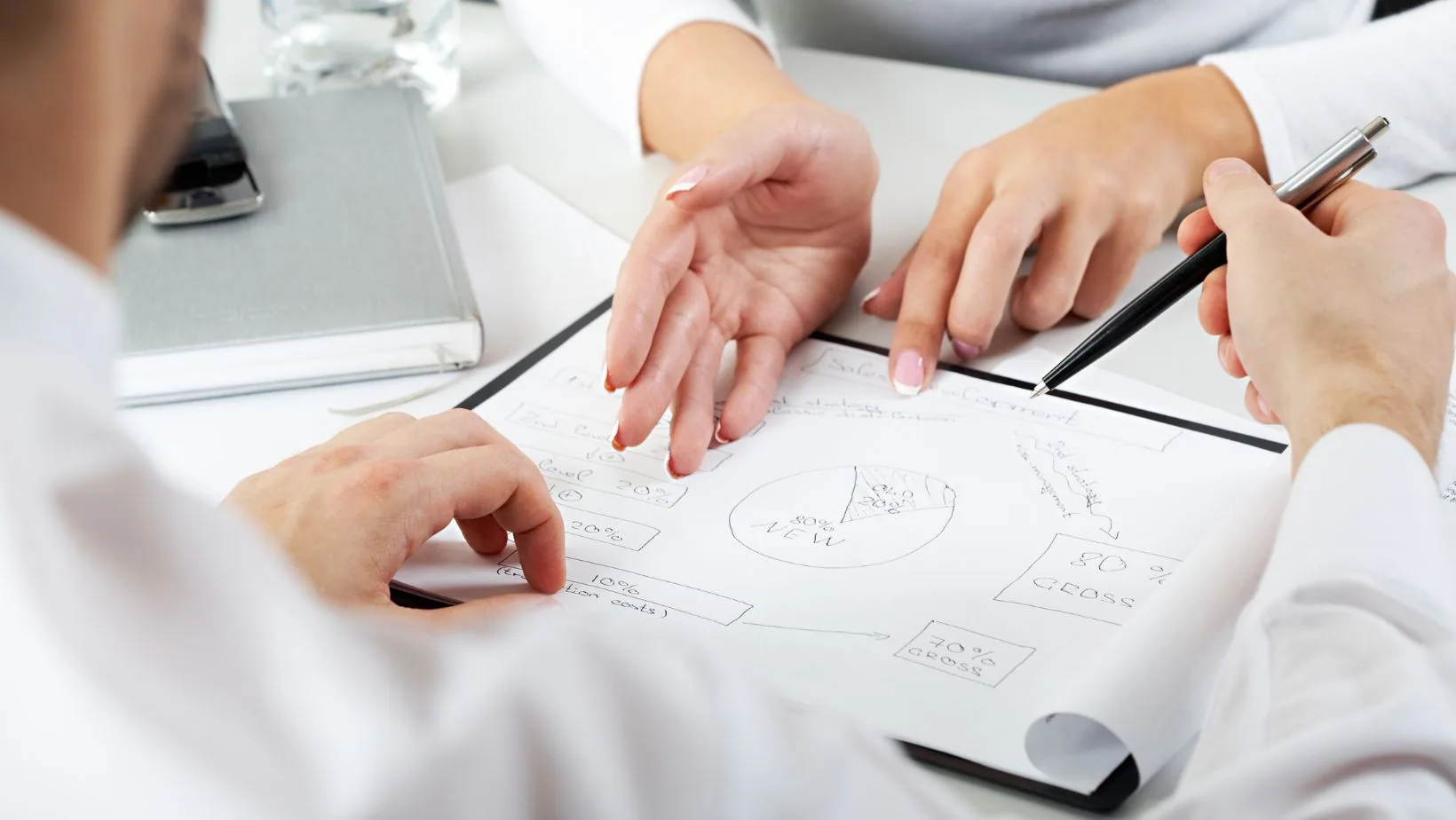 Multiple hands pointing to sketched diagrams and flowcharts on white paper during a collaborative discussion at a desk.