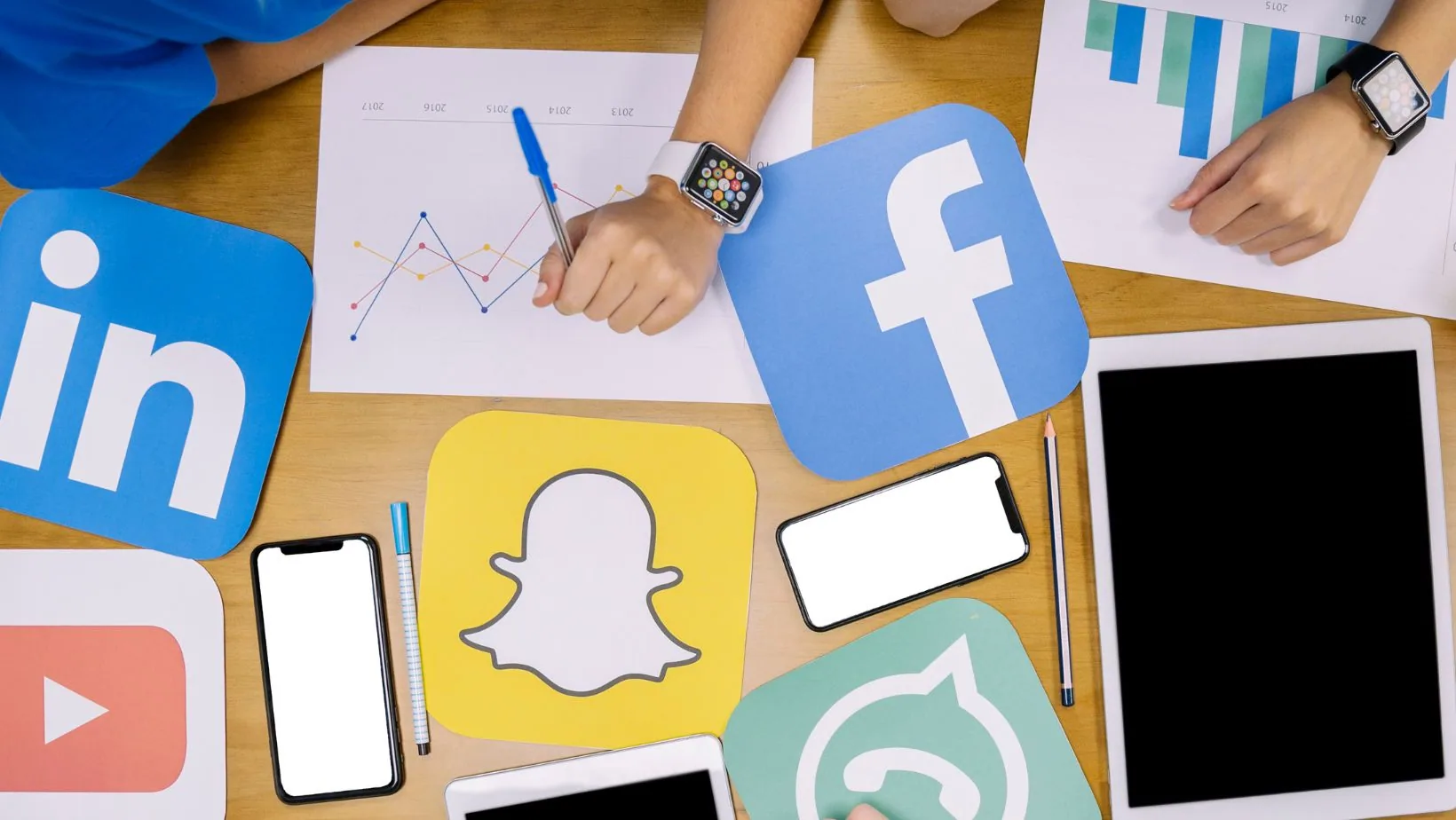 Hands holding pen over analytics charts surrounded by social media platform logos including Facebook, LinkedIn, Snapchat, and YouTube on wooden desk