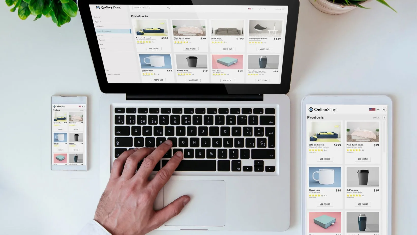 Hand resting on laptop keyboard displaying online shop product grid, with smartphone and tablet showing same e-commerce interface on white desk