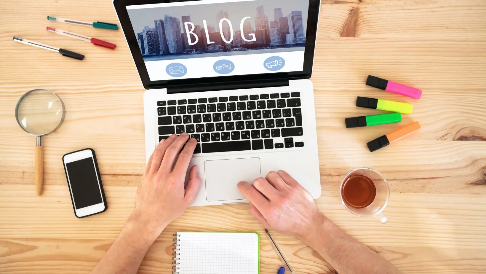 Person's hands typing on laptop displaying blog website with cityscape background, surrounded by office supplies on wooden desk