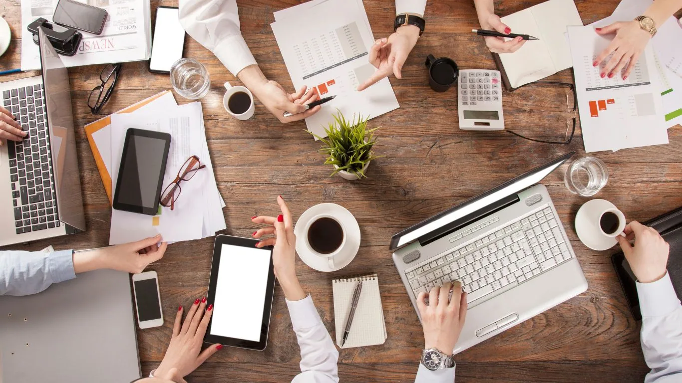 Overhead view of team members collaborating at wooden table with laptops, tablets, coffee cups, and business documents