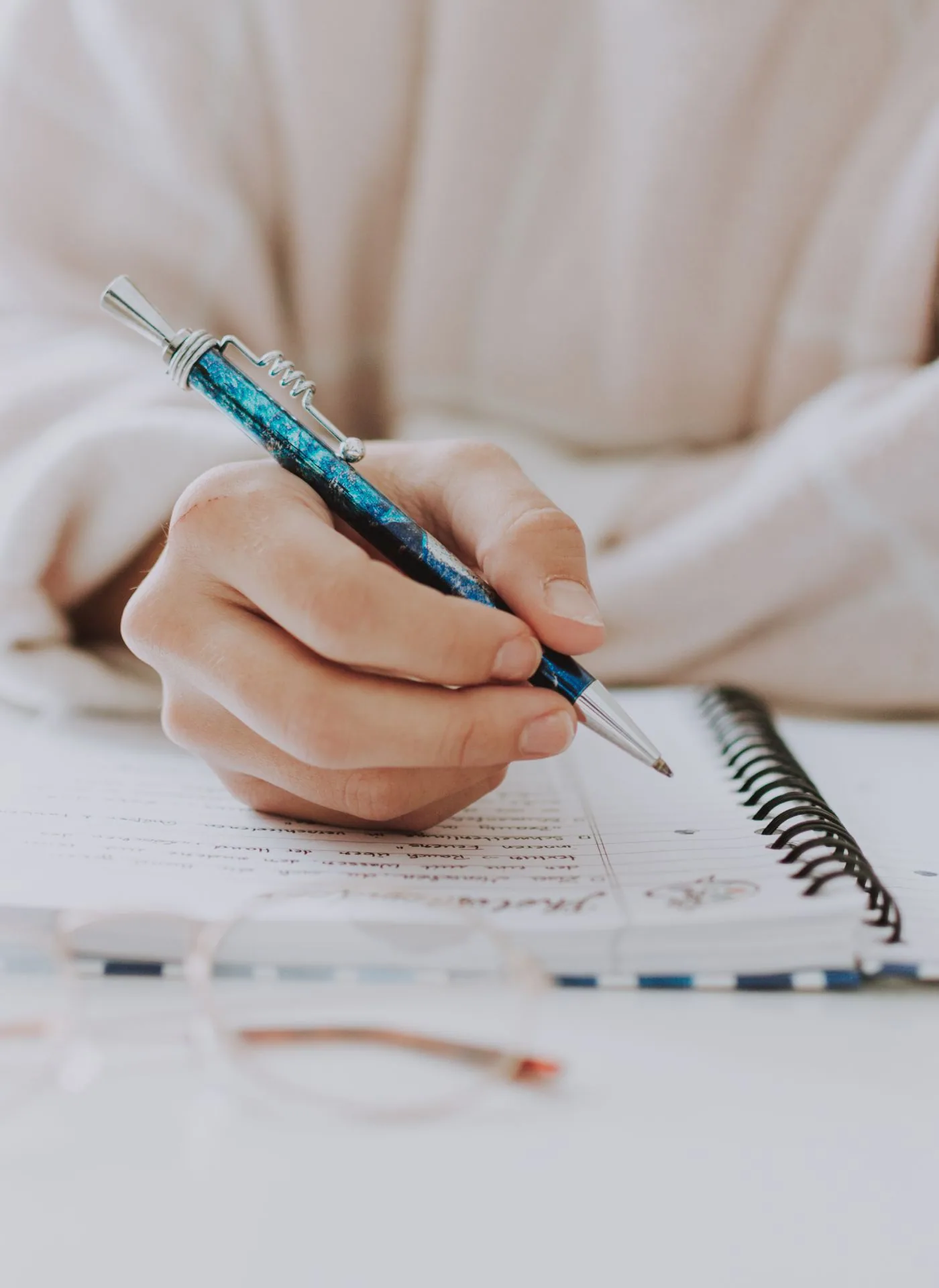 Hand holding a teal and silver pen poised over an open spiral notebook on a white desk