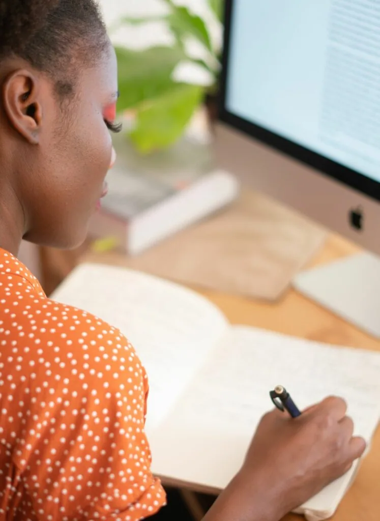Person in orange polka dot shirt holding pen at desk with notebook and computer monitor